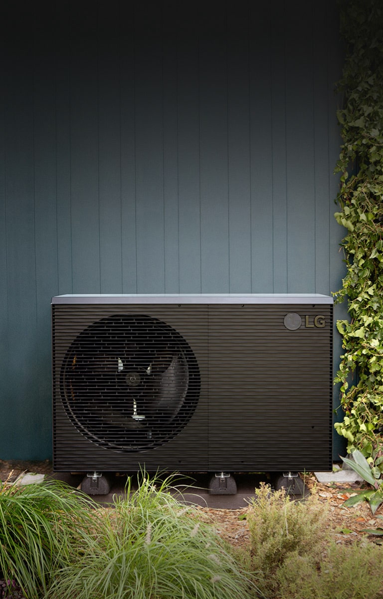 LG Air to Water Heat Pump THERMA V R290 Monobloc, black-colored outdoor unit is placed on the exterior green wall of the house.