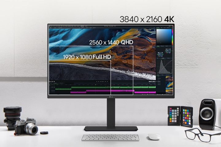 Front view of a black LG monitor on a desk, displaying layered screen resolutions for comparison, with cameras, books, colour chips, and speakers arranged around it.