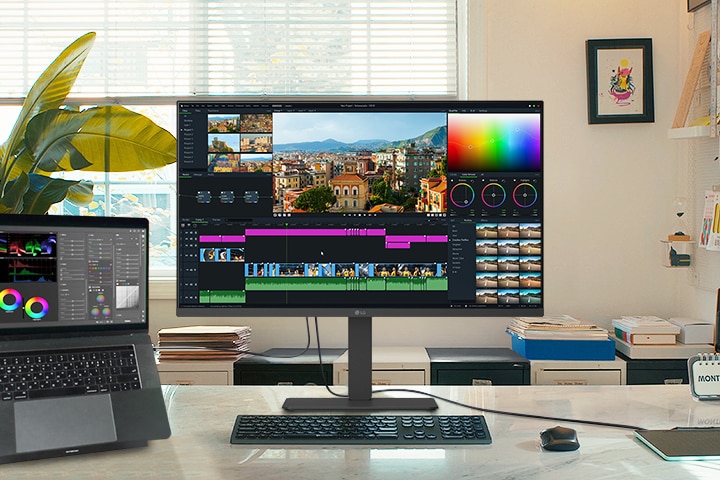 A creative workspace featuring an LG monitor and a connected laptop, both displaying a video editing interface with colour grading tools and a timeline.