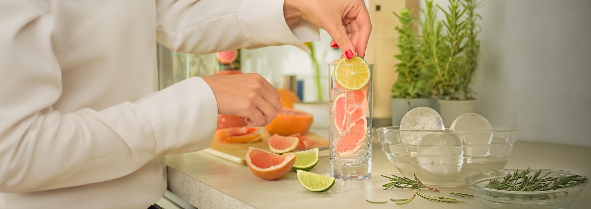 A woman putting grapefruits and a lime inside a tall glass, fruits, herbs and a bowl of ices are scattered around the glass.