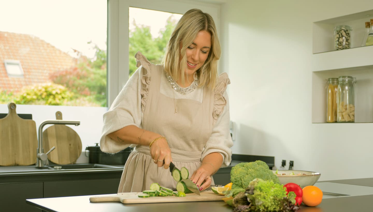 A woman chopping fresh cucumbers in a bright kitchen with a variety of colorful vegetables on the counter.