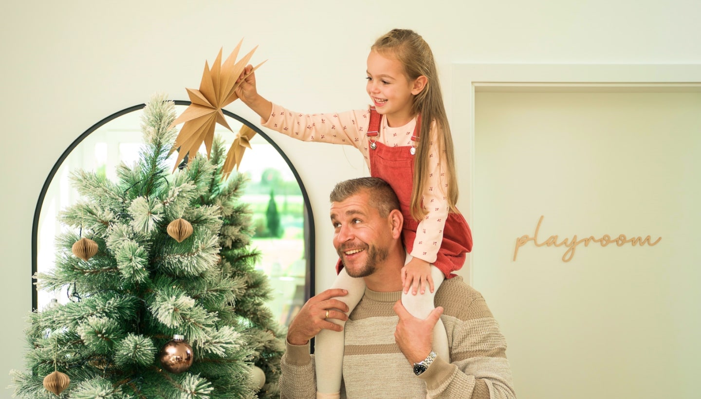 A father lifts his smiling daughter on his shoulders as she decorates the Christmas tree. Nearby, a door labeled 'playroom' is visible.