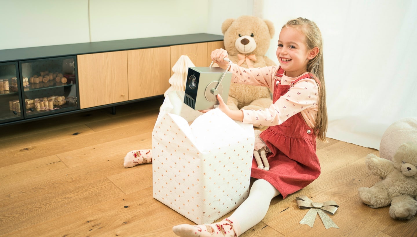 A smiling young girl opening a Christmas gift to reveal an LG CineBeam projector. She sits on a wooden floor.