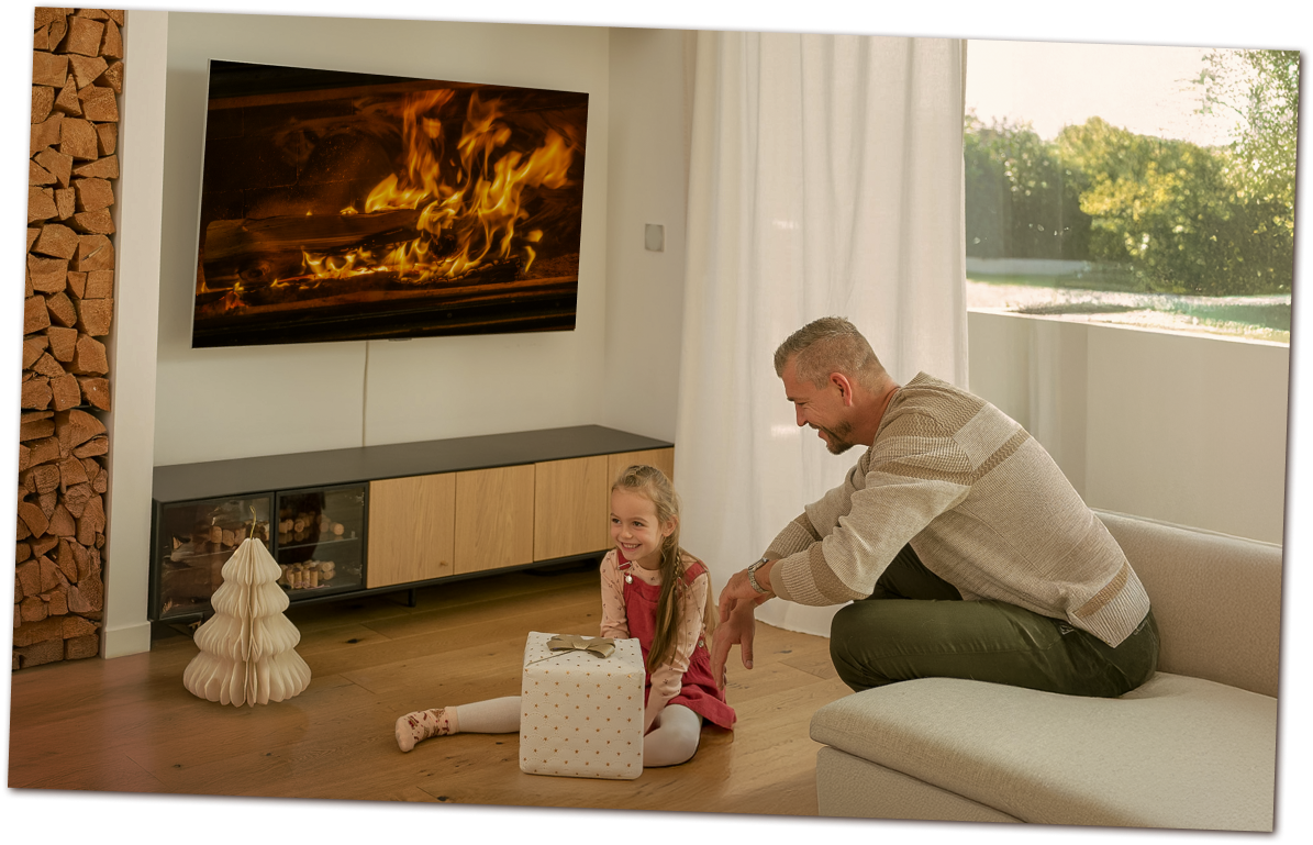 Father and daughter enjoying Christmas by an LG OLED TV displaying a warm fireplace. The girl holds a gift, surrounded by cozy décor.