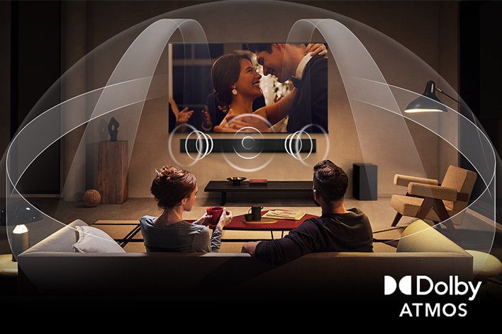 Dimly lit, cozy living room with a couple watching a movie on their LG OLED TV with LG Soundbar. Graphics show how Dolby Atmos fills the whole space with surround sound from all directions. Dolby Atmos logo is at the bottom corner.	