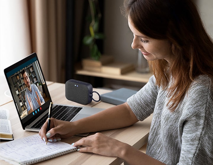 A woman in a video conference call with a co-worker, writing down on a note at the same time. XBOOM Go XG1 speaker is placed on a desk.