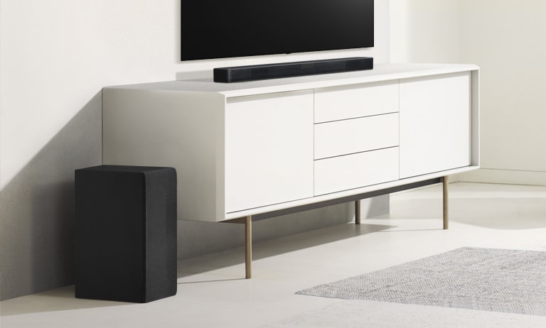 Loa Sound bar trên kệ màu trắng và bên trái có loa siêu trầm. Loa siêu trầm trông lớn hơn vì loa hướng về phía trước nhiều hơn.