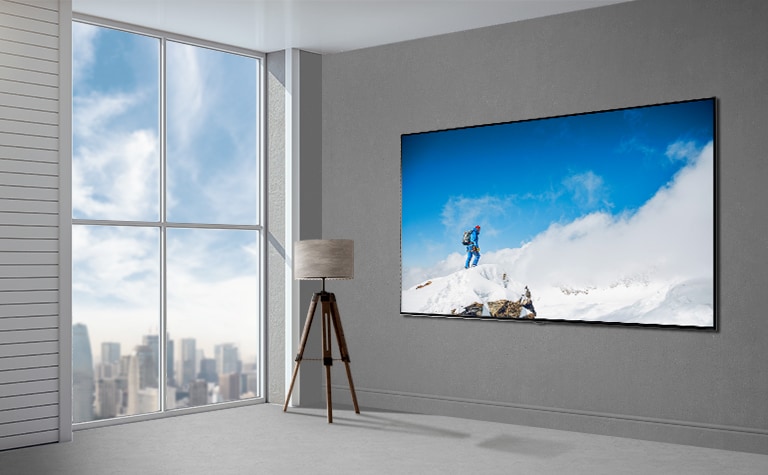 Hình ảnh một chiếc TV màn hình phẳng gắn trên tường màu xám cạnh cửa kính cao từ sàn đến trần nhìn ra toàn cảnh thành phố. Một chiếc đèn đặt bên trái TV và màn hình hiển thị hình ảnh một người đi leo núi trên ngọn núi phủ đầy tuyết trong nền trời xanh mây trắng.