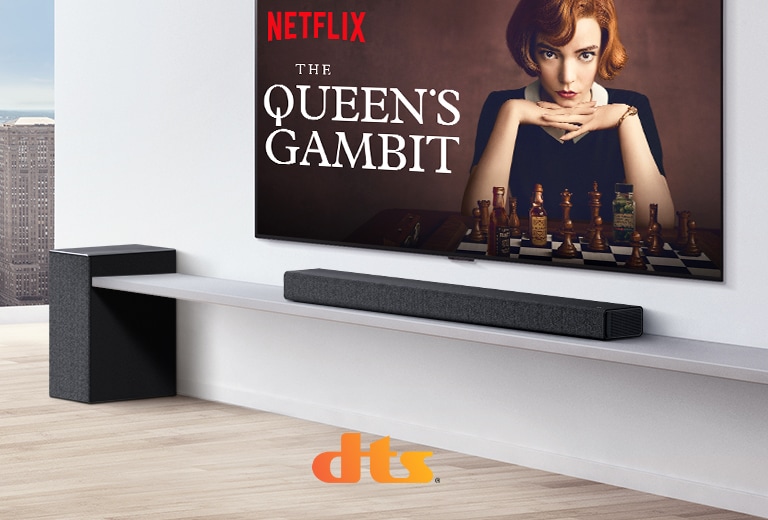 TV treo trên tường. Áp phích của chương trình truyền hình trên màn hình TV. LG Soundbar nằm ngay bên dưới TV trên kệ màu trắng với loa siêu trầm ngay bên cạnh. Logo Dolby Atmos và DTS:X được hiển thị ở chính giữa bên dưới của hình ảnh.