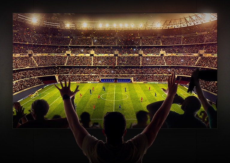 Four people sitting on a couch watching a large screen TV displaying a soccer stadium filled with spectators.