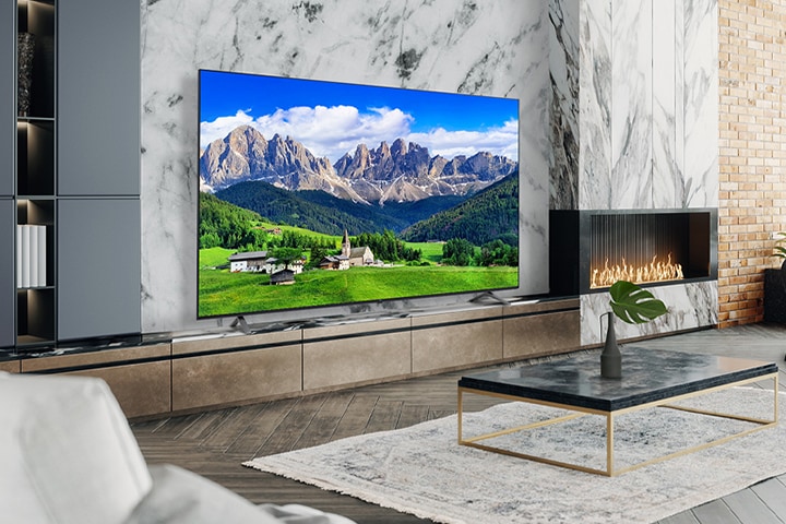 TV lớn treo trên tường phòng khách với lò sưởi trang trí. Cảnh núi non và làng mạc hiện lên sáng rõ và sống động trên màn hình TV.