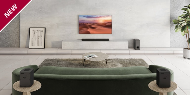 TV is hung on the white pillar in the center of the living room. Below, LG Sound Bar is placed on the white shelf. On the floor right under the sound bar, a wireless subwoofer stands. Behind the green sofa, two rear speakers are placed on the coffee table both the left and right sides. NEW mark is shown in the top left corner.