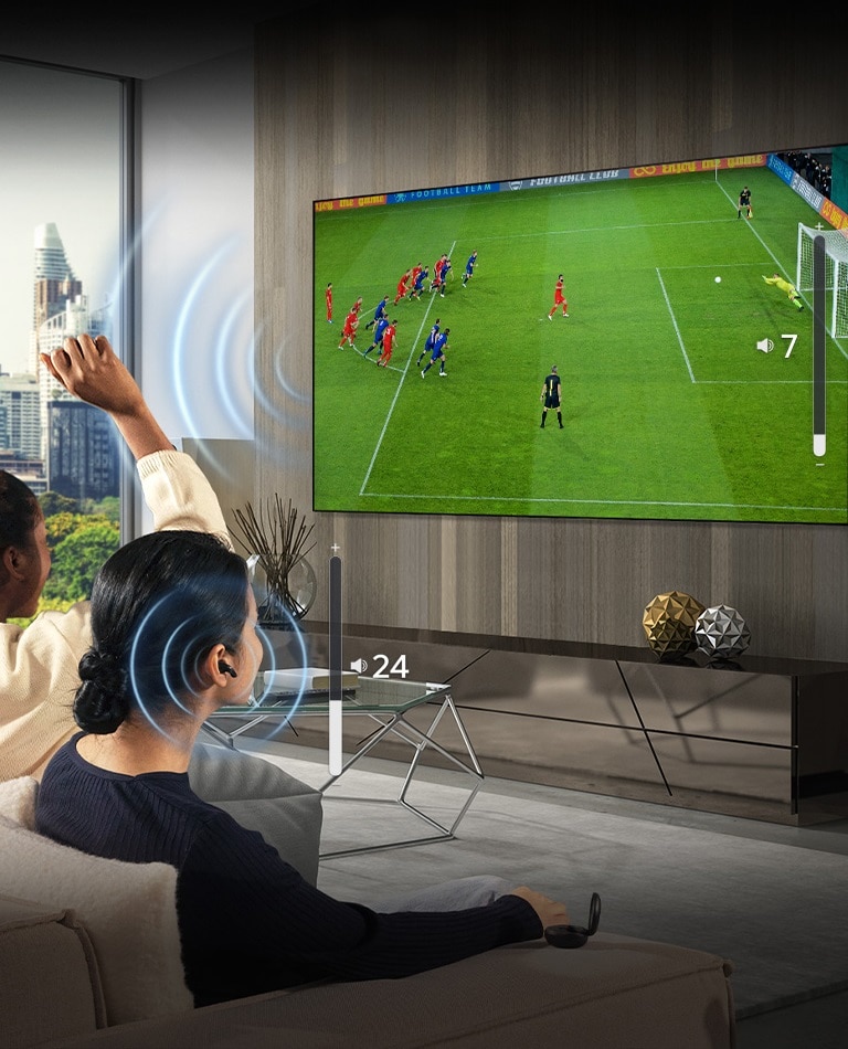 alt="A group of people are sitting on a sofa watching a soccer game on TV. The woman on the far right is wearing earbuds and using them with a different volume than the TV, indicating that she is using both at the same time."