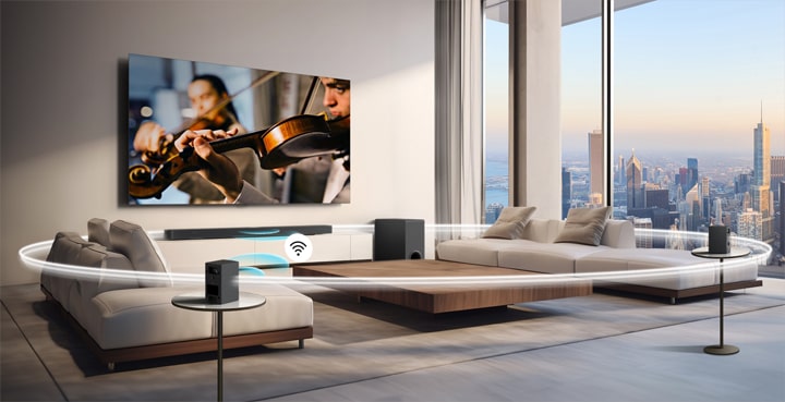 LG TV, LG Soundbar, a subwoofer, and rear speakers in a modern apartment overlooking the city. The rear speakers sit on minimalist side tables behind two sofas. A white circular band denoting connectivity stretches from the soundbar to subwoofer and rear speakers. Blue waves appear from soundbar and one rear speaker in left and a connectivity symbol bounce between the soundbar to the rear speaker. The TV shows two people playing the violin. 