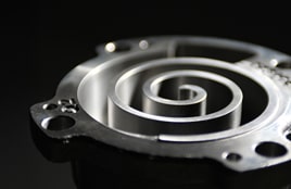 Nahaufnahme eines metallischen Spiralverdichterbauteils mit einer glatten, polierten Oberfläche vor dunklem Hintergrund.