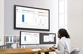 Eine Frau arbeitet an einem Schreibtisch in einem Büro und analysiert Daten, die auf mehreren Monitoren, darunter einem großen an der Wand montierten Bildschirm, angezeigt werden.