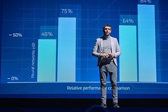 Professional speaker presenting on stage in front of a large digital screen with bar graphs comparing neural network performance percentages