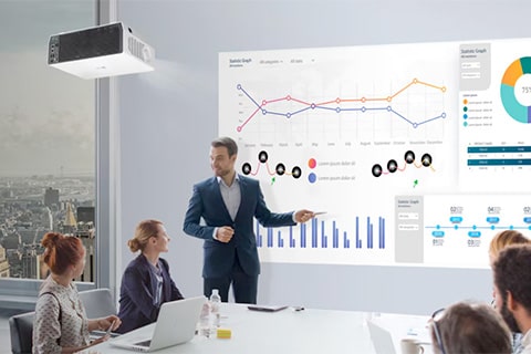 Business meeting scene where a man in a suit presents charts, graphs, and data visualizations on a large display screen to seated colleagues