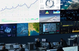 Control room with multiple large display screens showing data charts, maps, and monitoring dashboards while people work at computer stations.