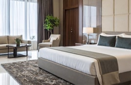 Modern hotel bedroom interior featuring a neatly made bed, soft lighting, armchairs, and large windows with natural light.
