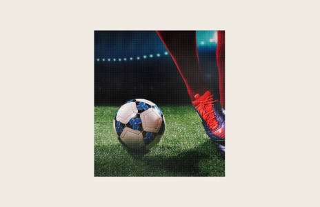 Close-up of a footballer wearing red boots beside a football on an artificial pitch under stadium floodlights.