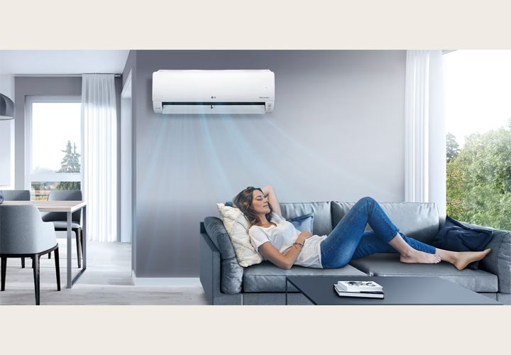 Mulher relaxa em um sofá enquanto um ar-condicionado LG na parede refrigera o ambiente, criando uma atmosfera confortável e agradável.