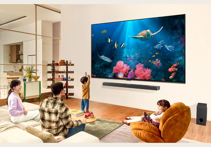 Uma família em uma sala de estar assiste a uma cena debaixo d’água com corais vibrantes e criaturas marinhas em uma grande TV LG colocada na parede. Abaixo da TV LG, uma elegante soundbar LG com subwoofer complementa a experiência na sala espaçosa e moderna.