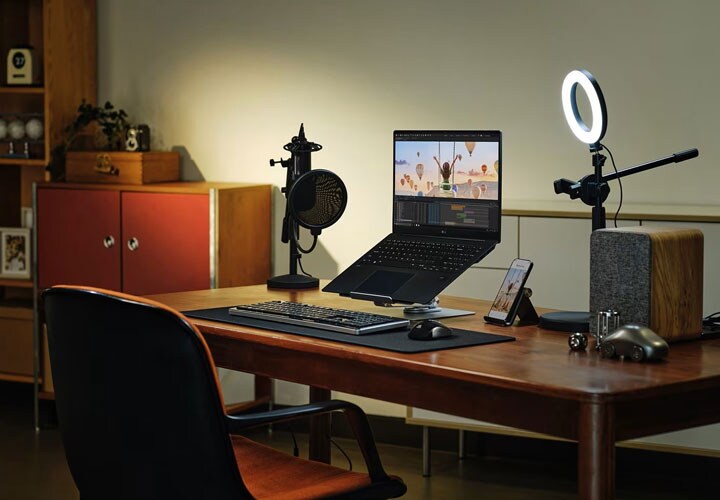 Uma configuração de home office com um laptop LG gram, teclado, mouse, microfone e ring light em uma mesa de madeira. Prateleiras e um armário estão ao fundo