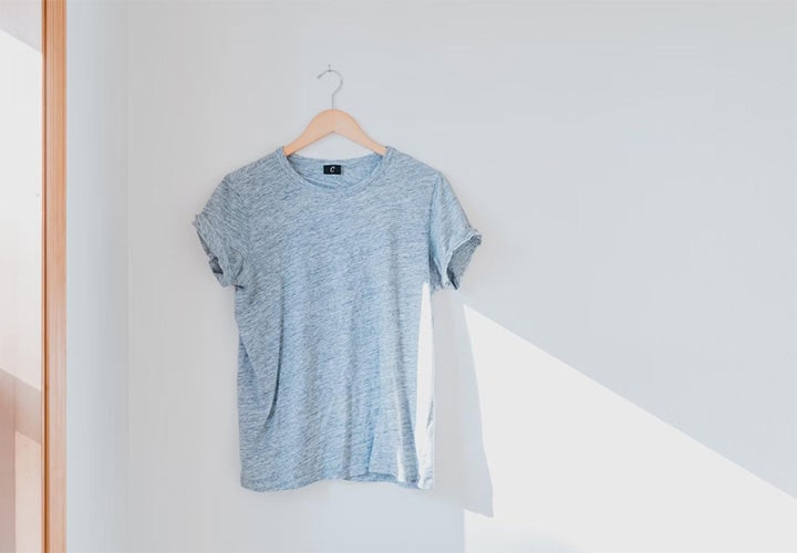 Uma camiseta azul-claro está pendurada em um cabide diante de uma parede branca.
