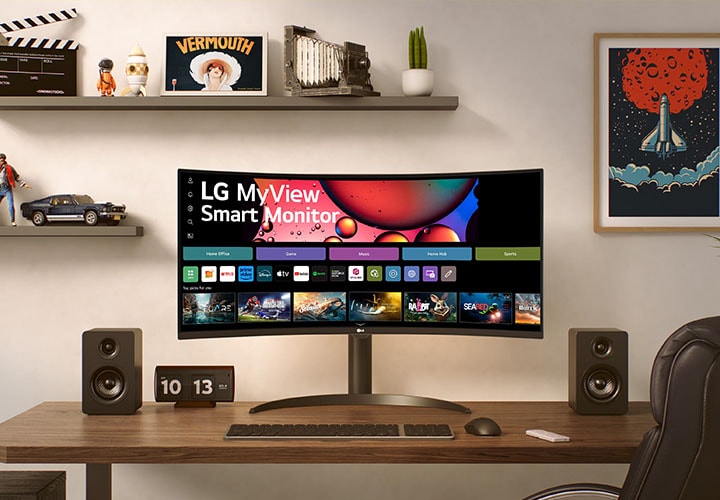 Um monitor smart LG em uma mesa moderna com caixas de som e um relógio. No fundo, prateleiras com decoração e arte na parede, deixando o espaço de trabalho com muito estilo.