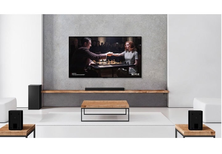  Uma sala de TV minimalista com um conjunto de soundbar e subwoofer e uma TV LG instalada na parede. A tela da TV exibe um título da Netflix, com duas caixas de som traseiras que oferecem um som imersivo.