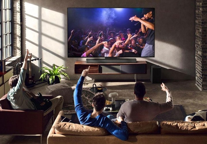Texto alternativo: Em uma sala em estilo loft, um grupo de amigos assiste a um show em uma TV de tela grande pareada a uma soundbar LG com subwoofer. O ambiente é aconchegante e vibrante, destacando a experiência de som surround.