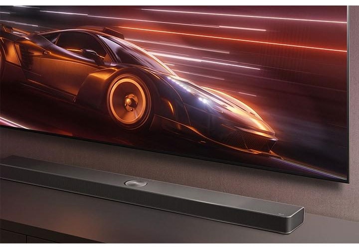 Texto alternativo: Imagem aproximada de uma soundbar LG colocada em um rack de TV enquanto um dinâmico jogo de corrida é exibido, enfatizando a experiência de jogo imersiva.