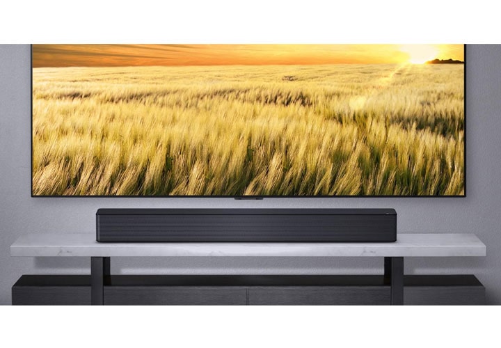 Texto alternativo: Uma soundbar LG posicionada abaixo de uma TV de tela grande que exibe um campo de trigo. O elegante rack de mármore complementa o design refinado da soundbar.