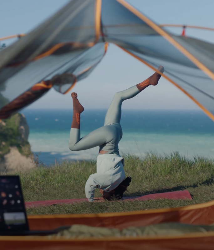 Une femme faisant le poirier sur un tapis de yoga au bord de l'océan, filmant avec son smartphone affiché sur un LG Gram.