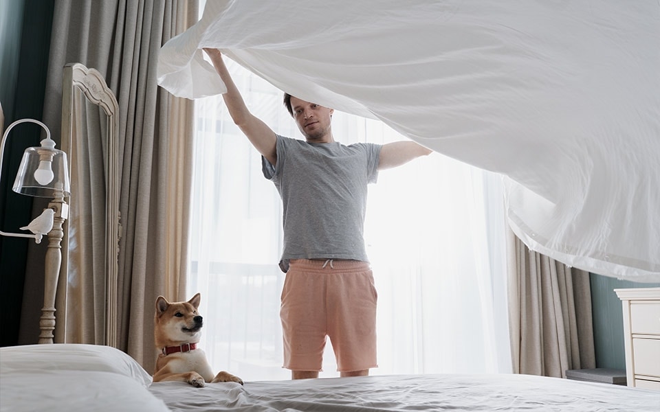 A man places a white sheet on his bed with his dog standing next to him