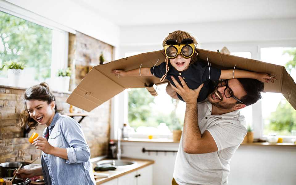 A child wearing cardboard wings being lifted in the air by a happy man in the kitchen.