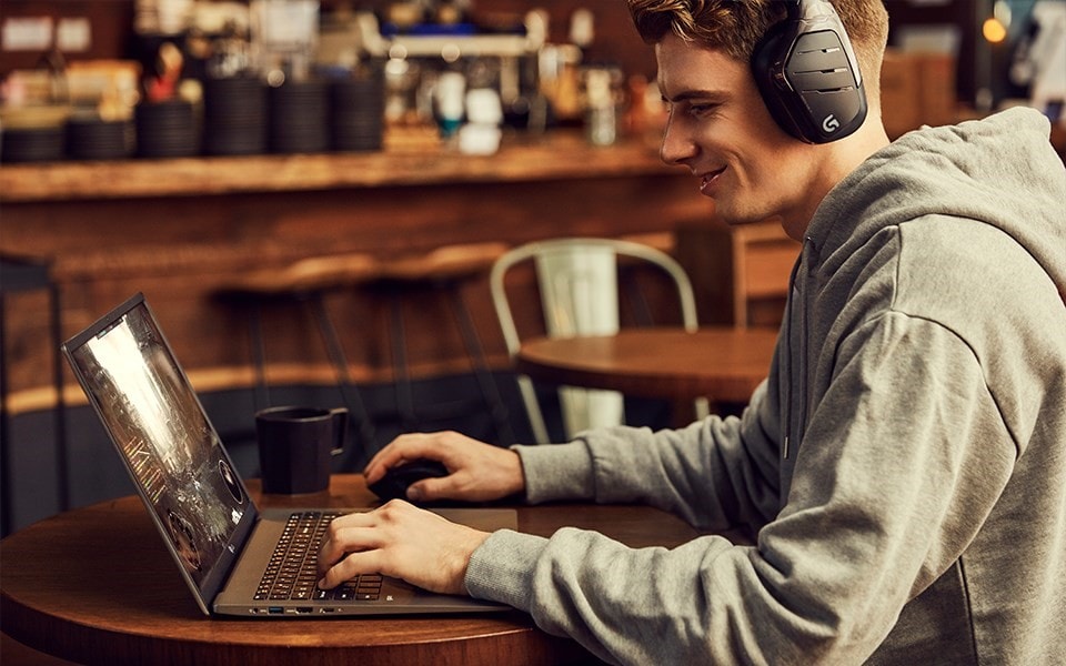 A young man games on a laptop in a coffee shop with headphones on