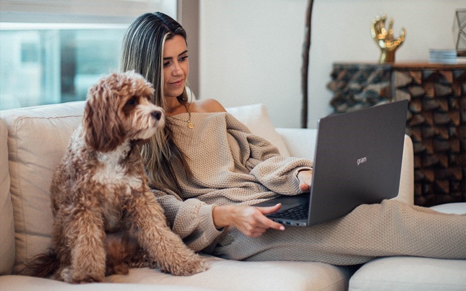 A woman sits on a sofa with a laptop on her lap and her dog beside her