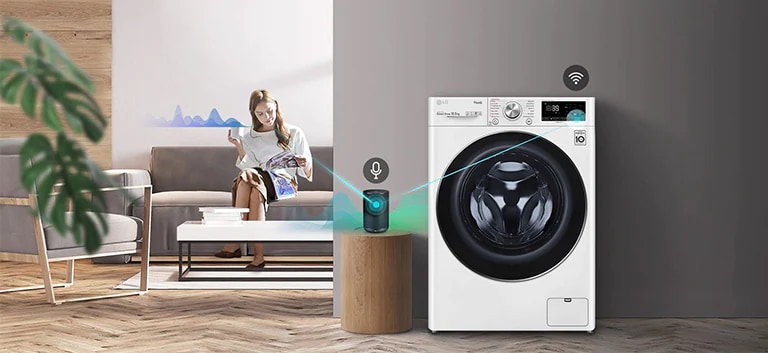 A woman sitting on the couch and drinking tea is giving orders to her washing machine through her AI speaker.