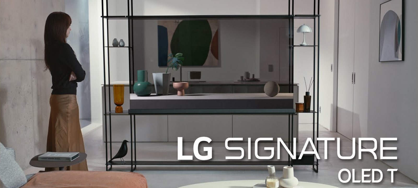 A person stands in a modern living room next to an LG SIGNATURE OLED T with transparent screen, with decorative items and abstract art in the background.