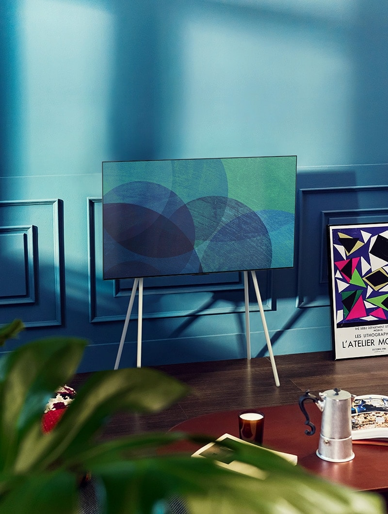 Een LG OLED televisie op Gallery stand staat in een moderne grijze kamer voor een raam dat uitkijkt op een besneeuwd bos. Een LG OLED televisie met draaistandaard staat in een kleurrijke woonkamer met pastelkleuren en houten meubilair.