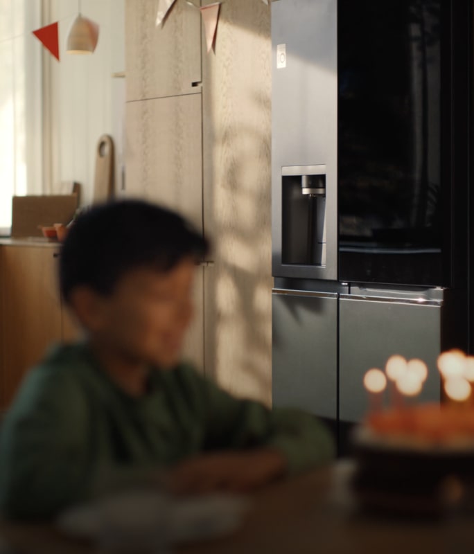 The child's face is faintly visible in front of the candlelit cake, and the LG InstaView refrigerator is clearly visible in the background.