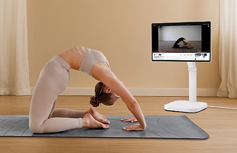 Kvinna i sportkläder utför en bakåtböjning på en yogamatta inomhus. Hon använder en LG Swing, en vit skärm på ett stativ, placerad framför henne.