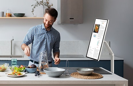 En man lagar mat i ett modernt kök samtidigt som han använder en LG Smart Monitor monterad på en Swing-arm på köksbänken. Skärmen är vinklad mot honom och visar innehåll medan han förbereder mat. Köket är ljust och minimalistiskt, med tallrikar och ingredienser på bänken