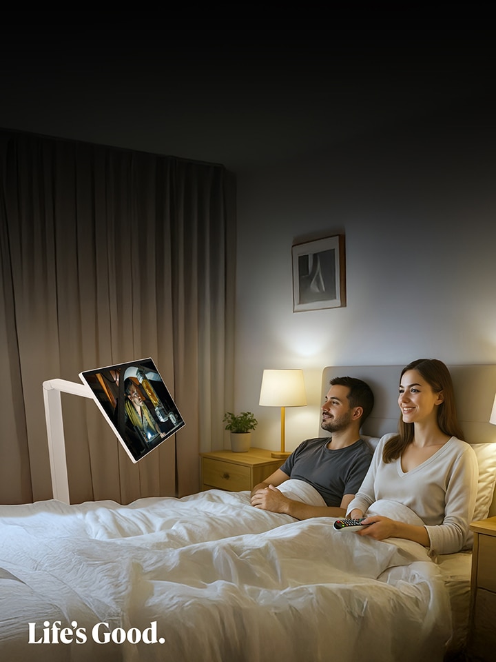 Ett par ligger i sängen och tittar på en LG Smart Monitor som är monterad på en Swing-arm framför dem. Rummet är mjukt upplyst med en sänglampa, och skärmen visar färgglatt innehåll. Båda ser avslappnade ut. Längst ner i bilden syns logotyperna för LG och Swing Smart Monitor.