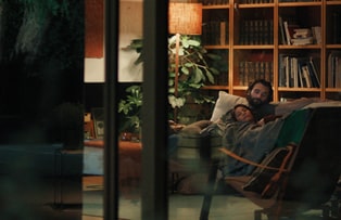A scene where the camera captures a man and a woman lying together on the living room sofa from a distance.