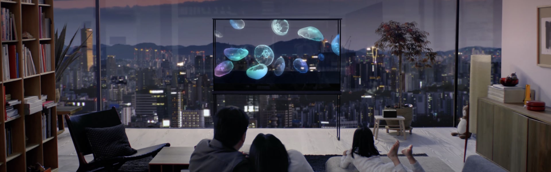 A family enjoys an LG Oled T with a jellyfish floating on it, while the cityscape shines through the transparent screen. 