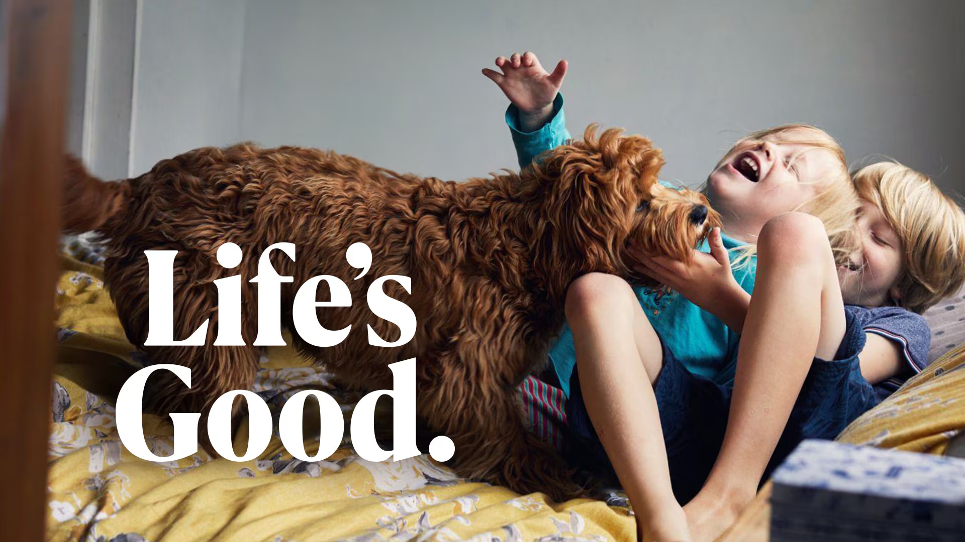 Two young boys are lying side by side on a bed, both with warm smiles on their faces, as they cuddle a friendly dog between them.