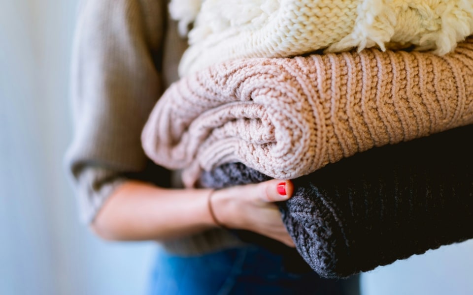 Eine Person hält einen Stapel gefalteter Pullover in verschiedenen Farben, darunter Cremeweiß, Rosa und Schwarz.